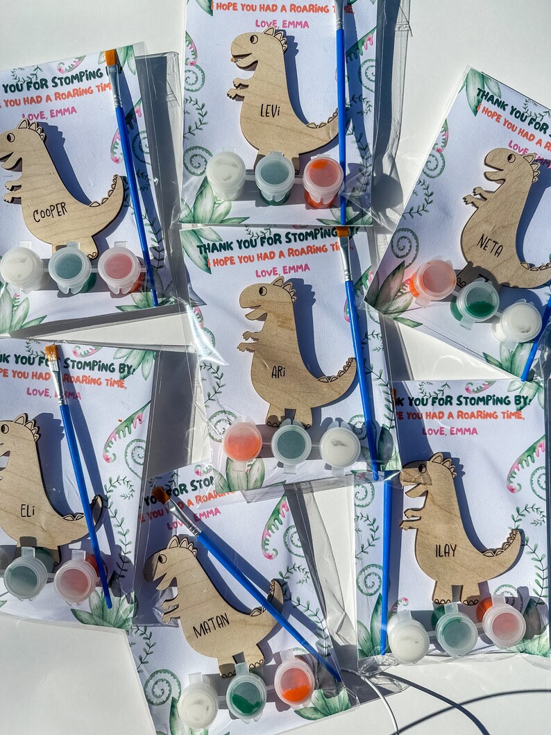 May include: A set of six paint-your-own dinosaur crafts, each with a different name. The dinosaurs are made of wood and have a light brown color. Each craft comes with three small bottles of paint, a paintbrush, and a card with the message "Thank you for stomping by! Hope you had a roaring time. Love, Emma".