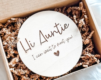 Op de afbeelding: Een houten cirkel met de tekst "Hi Auntie I can wait to meet you!" en een klein hartje in een bruine doos met gescheurd papier.
