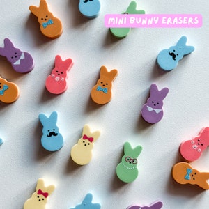 May include: A collection of colorful mini bunny erasers in various designs, including a blue bunny with a mustache, a pink bunny with flowers, and a green bunny with glasses. The erasers are arranged on a white surface with the text "MINI BUNNY ERASERS" in pink letters.
