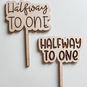 May include: Two wooden signs with the text "Halfway to One" written in black cursive. The signs are attached to wooden sticks.