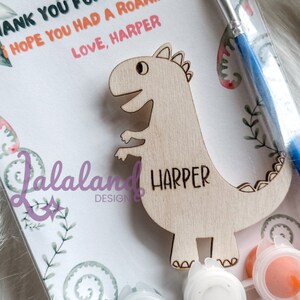 May include: A wooden dinosaur cutout with the name "Harper" painted on it. The dinosaur is accompanied by a paintbrush and three small paint pots with white, orange, and blue paint. The card reads "Thank you for stomping by. I hope you had a roaring time. Love, Harper."