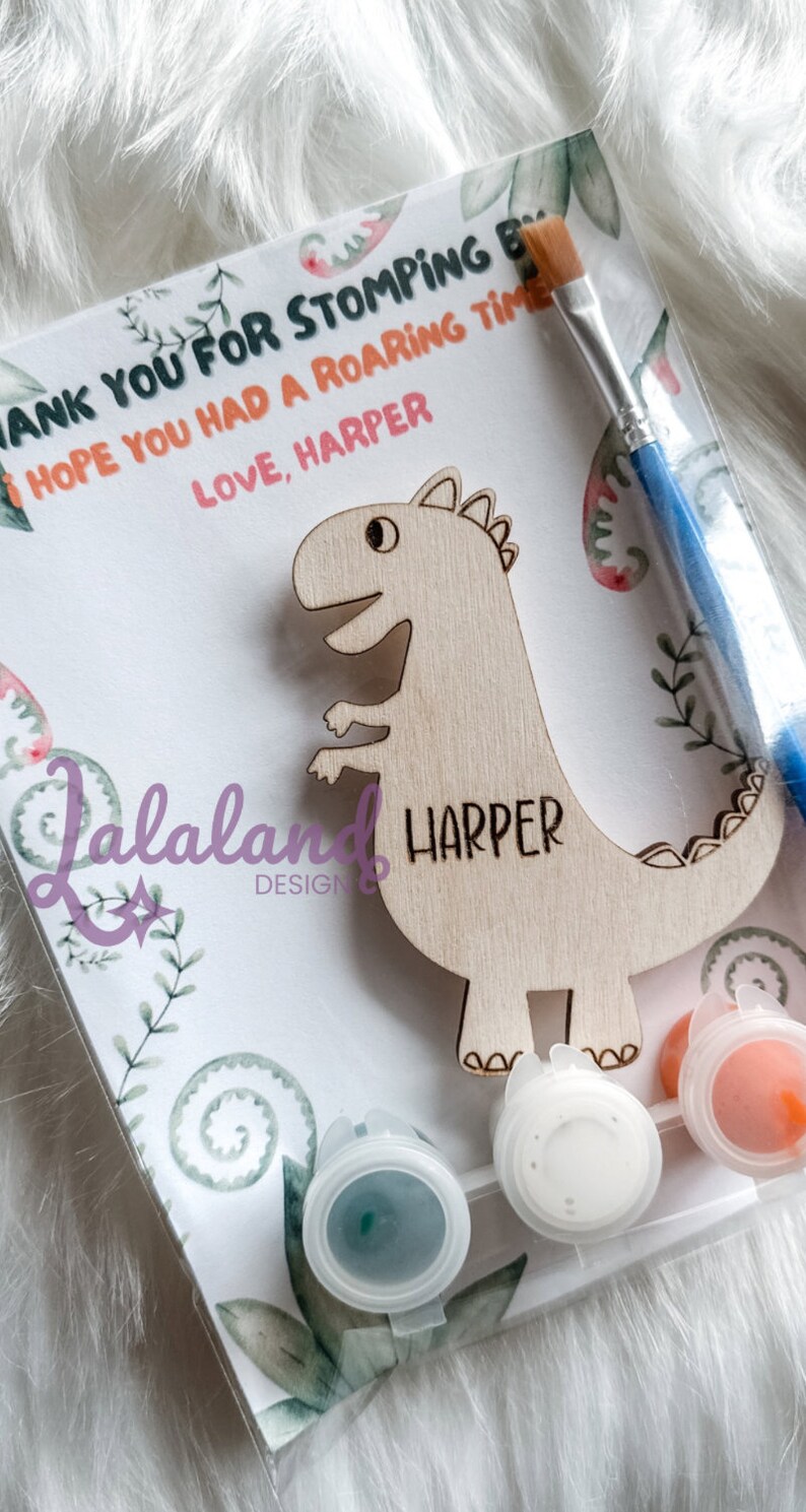 May include: A wooden dinosaur cutout with the name "Harper" painted on it. The dinosaur is accompanied by a paintbrush and three small paint pots with white, orange, and blue paint. The card reads "Thank you for stomping by. I hope you had a roaring time. Love, Harper."