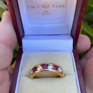 Könnte beinhalten: Ein goldener Ring mit abwechselnd roten und klaren Steinen, präsentiert in einer offenen, bordeauxfarbenen Schmuckschatulle. Der Ring steht im Mittelpunkt und zeigt sein Design. Auf der Schatulle steht "PERCY MARKS SYDNEY".
