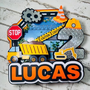 Puede incluir: Un adorno para pastel con temática de construcción con un número 2 plateado, un camión volquete amarillo, una excavadora amarilla, una señal de alto, un cono de tráfico, un casco y el nombre Lucas en letras naranja y negras.