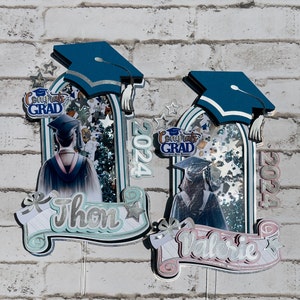 Puede incluir: Dos adornos para pasteles con temática de graduación con gorras azules, purpurina y los nombres "Thon" y "Valerie" en rollos. Los adornos presentan el año "2024" y la frase "Congrats Grad".