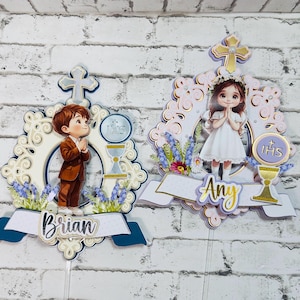 Pode incluir: Duas decorações de bolo de Primeira Comunhão. Uma apresenta um menino de fato castanho, a outra uma menina de vestido branco. Ambas têm detalhes florais, cruzes e os nomes "Brian" e "Any".
