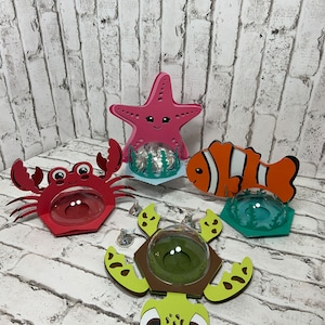May include: Four colorful cardboard boxes shaped like a starfish, crab, clownfish, and sea turtle. Each box has a clear plastic dome on top and is designed to hold small treats or favors.