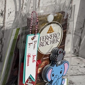 Puede incluir: Una bolsa de regalo de plástico transparente con un patrón de rayas verdes y blancas. La bolsa contiene una barra de chocolate, una pequeña bolsa de caramelos de chocolate y una etiqueta que dice "De: Jack A: Alanna". También hay un elefante azul con un ratón rosa encima.
