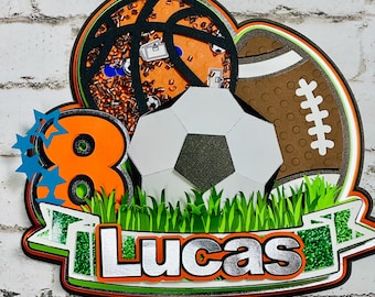 Puede incluir: Un adorno para pastel con un balón de fútbol, un balón de fútbol americano y un balón de baloncesto con el número 8 y el nombre Lucas en una pancarta de hierba verde.