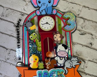 May include: A colorful, layered cake topper featuring a clock, animals, and the number 3. The topper includes a blue elephant, a mouse with cheese, a snake, a monkey, a duck, a frog, a squirrel, a cow, and a cat. The name "JACK" is displayed on a banner.