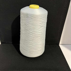 May include: A large spool of white thread with a yellow center. The thread is tightly wound and appears to be made of a smooth, durable material. The spool is set against a black background.