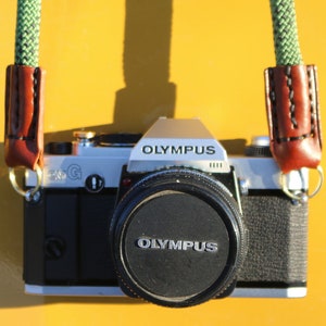 Op de afbeelding: Een zilveren en zwarte Olympus filmcamera met een groene en bruine leren band. De camera lens heeft het woord "Olympus" erop gedrukt.