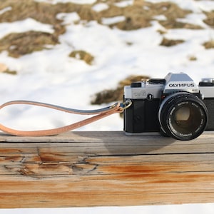May include: A silver Olympus OM-1 camera with a black lens and a brown leather strap. The camera is sitting on a wooden surface.