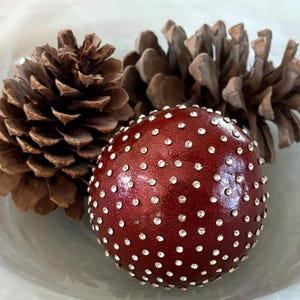 May include: A decorative burgundy sphere adorned with numerous small, sparkling rhinestones. Two brown pine cones are positioned behind the sphere. The items are arranged in a light-colored, shallow bowl, creating a festive, seasonal display.