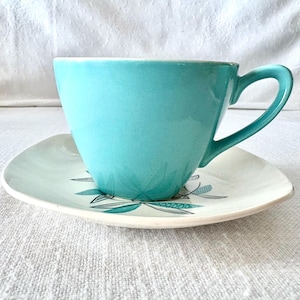 May include: A turquoise teacup and saucer set. The cup has a curved handle and a smooth, glossy finish. The saucer is square with rounded edges and a floral design in teal and gray.