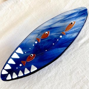 May include: An elongated, oval-shaped glass dish featuring a swirling blue and white marbled pattern. The design includes three small orange fish with black eyes and a series of white triangular shapes along one edge, resembling shark teeth.