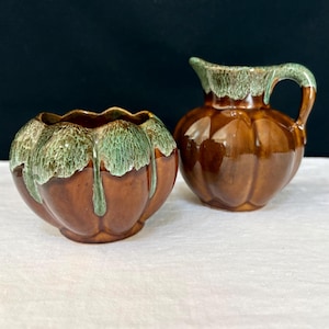 May include: A ceramic sugar bowl and a matching creamer pitcher, both featuring a glossy brown base with a scalloped, pumpkin-like shape and a decorative green drip glaze pattern cascading from the top rims down the sides of the vessels.