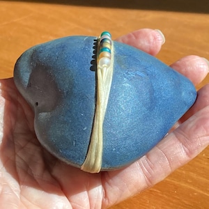 May include: A heart-shaped, blue stone with a cream-colored band wrapped around it. A row of colorful beads in shades of teal, gold, and white adorn the band. The stone is held in a hand, with a wooden surface in the background.