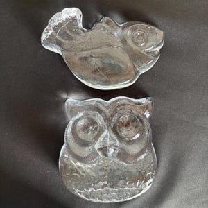 May include: Two clear glass decorative dishes shaped like animals. The top dish is shaped like a fish with a textured surface and a defined eye. The bottom dish is shaped like an owl with large circular eyes, a pointed beak, and textured feathers.