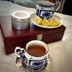 May include: A still life featuring a cup of tea, a small tin, and a decorative cup and saucer set. The cup and saucer have a blue and white floral design. Slices of lemon are on a white plate. The items are on a wooden box.