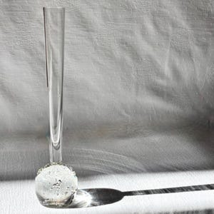 May include: A clear glass bud vase with a round, textured base and a long, slender neck. The base is decorated with small, bubble-like details. The vase casts a long shadow on a white, textured surface.
