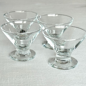 May include: Four clear glass dessert dishes. Each dish has a rounded bowl shape with a flared, conical base and a circular foot. The glass is transparent, allowing light to pass through, and the dishes are arranged on a neutral surface.
