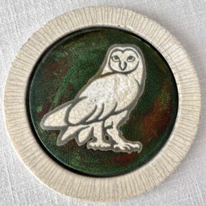 May include: A round ceramic coaster featuring a white owl design on a dark green background. The owl is outlined in gray and has a cracked, aged appearance. The coaster has a textured, off-white border.