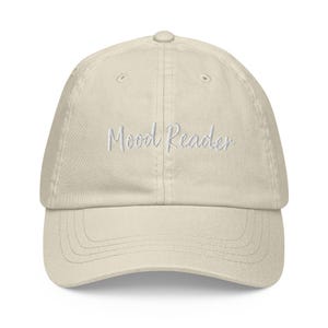 May include: A beige baseball cap with a six-panel construction and a curved brim. The front of the cap features the text "Mood Reader" embroidered in white cursive script. The hat includes a button at the top and stitched eyelets for ventilation.