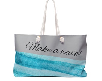 Könnte beinhalten: Eine graue Canvas-Tragetasche mit weißen Kordelgriffen. Die Tasche hat ein blaues Aquarelldesign und den Text "Make a wave!" auf der Vorderseite.