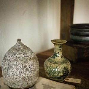 May include: Two ceramic vases on a wooden surface. One vase is a bulbous shape with a textured, speckled pattern. The other is a smaller, round vase with a green and blue floral design. An open book is in the foreground.