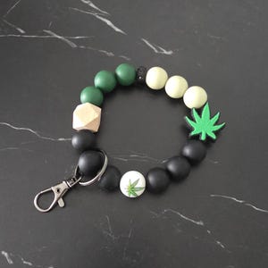 May include: A circular keyring bracelet featuring a mix of matte black, dark green, and light green silicone beads, a natural wood geometric bead, a bright green leaf-shaped charm, and a white bead with a small green leaf print, attached to a metal clasp.