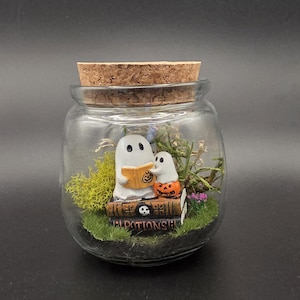 May include: A clear glass jar with a cork lid containing a miniature scene of two white ghosts sitting on stacked books. One ghost holds an orange book with a pumpkin design, while the other sits on a small orange pumpkin. Green moss and tiny flowers surround them.