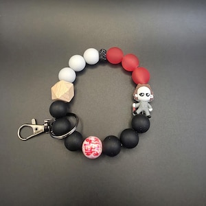 Op de afbeelding: Een ronde sleutelhangerarmband met matte zwarte, witte en rode siliconen kralen, een geometrische kraal van natuurlijk hout en een kleine zwarte glitterkraal. Het bevat een figuurbedel met een mes en een kraal met rode handafdrukken.