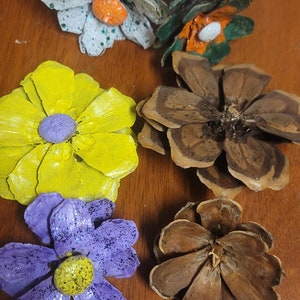 Może przedstawiać: Sześć szyszek sosnowych pomalowanych tak, aby wyglądały jak kwiaty. Kwiaty są pomalowane na zielono, biało, żółto, fioletowo i brązowo. Kwiaty mają pomarańczowe, żółte i fioletowe środki.