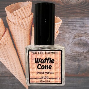Puede incluir: Un pequeño frasco de vidrio de perfume con aroma a cono de waffle. La botella está etiquetada como "Pixie Spell Essentials Waffle Cone Eau de Parfum" y enumera los ingredientes como "Perfumers Alcohol, Skin Safe Fragrance Oil" y el tamaño como "0.5 fl oz | 15ML".