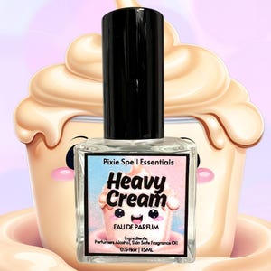 Puede incluir: Una botella de perfume de vidrio transparente con una tapa negra. La etiqueta es rosa y azul con el texto "Pixie Spell Essentials", "Heavy Cream", "Eau de Parfum", "Ingredientes: Alcohol de perfumería, Aceite de fragancia seguro para la piel", y "0.5 fl oz | 15ML".