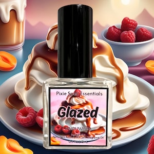 May include: A small glass bottle of perfume with a black cap. The label reads "Pixie Spell Essentials" and "Glazed" in pink letters. The bottle is sitting on a white plate with a dessert made of whipped cream, caramel sauce, and raspberries.