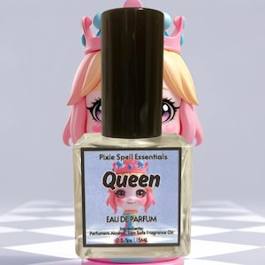 May include: A square perfume bottle with a black cap, labeled "Queen" with a cartoon image. The bottle is in front of a pink and yellow figurine. The label reads "Pixie Spell Essentials" and "EAU DE PARFUM".