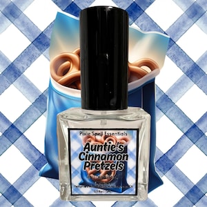 Puede incluir: Una botella de vidrio transparente con una tapa negra y una etiqueta blanca. La etiqueta dice "Pixie Spell Essentials Auntie's Cinnamon Pretzels Eau de Parfum Perfume Oil 0.5 fl oz | 15 ml". La botella está sobre un fondo a cuadros azules y blancos.
