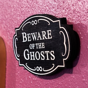 May include: Black wooden sign with white lettering that reads "Beware of the Ghosts". The sign has a decorative border with an infinity symbol.