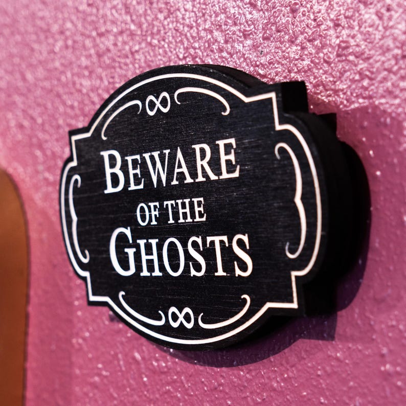 May include: Black wooden sign with white lettering that reads "Beware of the Ghosts". The sign has a decorative border with an infinity symbol.