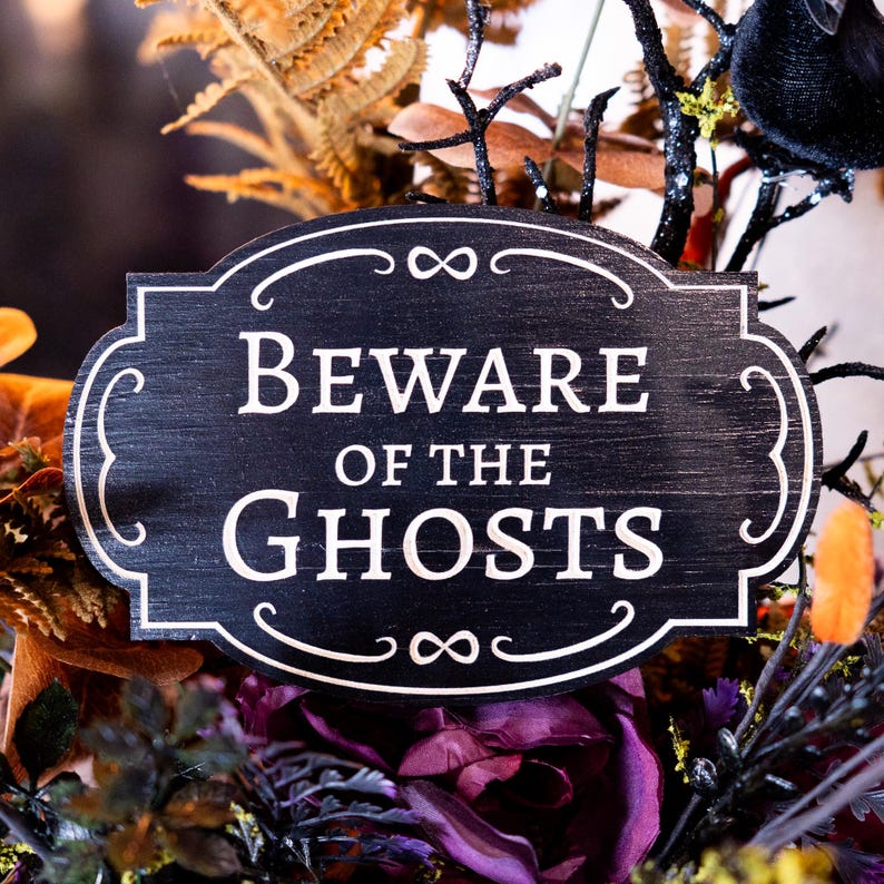 May include: A black wooden sign with white lettering that reads "Beware of the Ghosts". The sign is decorated with an infinity symbol on either side.