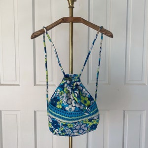 Può includere: Uno zaino con coulisse blu e verde a fiori su sfondo bianco. Lo zaino è appeso a un appendiabiti in legno con un pomo a testa di cavallo dorato.