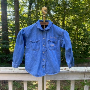 Pode incluir: Camisa jeans azul claro com mangas compridas, dois bolsos frontais e gola com babados. A camisa apresenta detalhes florais bordados e o texto "My Sunshine".