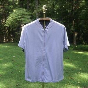 Puede incluir: Una camisa azul claro de manga corta con cremallera y rayas blancas en los hombros. La camisa está colgada de una percha de latón sobre un fondo de hierba verde y árboles. La camisa tiene una cremallera completa.