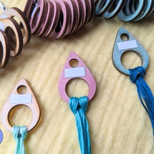 May include: Three wooden thread holders in different colours, pink, light blue, and natural wood. Each holder has a small ring and a white label. The holders are attached to strands of embroidery floss in different colours.