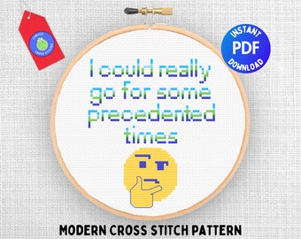 May include: A cross stitch pattern featuring a pixelated design of a thinking face emoji and the text "I could really go for some precedented times". The pattern is displayed in a wooden embroidery hoop on a white fabric background.