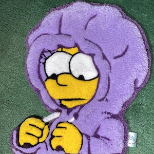 May include: A purple rug featuring a cartoon character in a hooded sweatshirt. The character has yellow skin, large white eyes, and yellow hands. The rug is placed on a green surface.