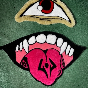May include: Two tufted rugs featuring graphic designs. The top rug is an eye with a red iris, white sclera, and tan border. The bottom rug is an open mouth with white teeth, pink gums, and a bright pink tongue displaying a black symbol in the center.