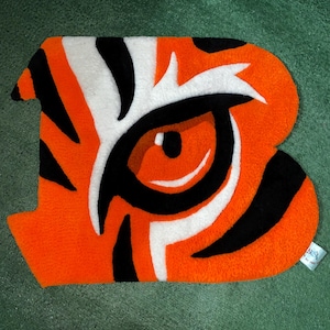 May include: Orange, black, and white rug in the shape of a letter "B" with a tiger eye design. The rug has a plush texture and is placed on a green surface. The eye is detailed with black and white accents.